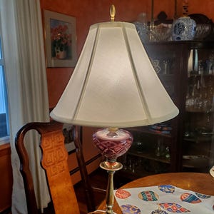 May include: A gold table lamp with a white lampshade and a burgundy glass base. The lamp is sitting on a table with a patterned tablecloth.