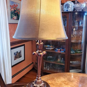 Könnte beinhalten: Eine goldene Tischlampe mit einem braunen Lampenschirm. Die Lampe hat einen dekorativen Sockel mit einem floralen Design. Die Lampe steht auf einem Holztisch.