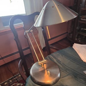 May include: A brushed metal desk lamp with a conical shade and a round base. The lamp has a minimalist design with a pivoting arm and a small on/off switch. The lamp is positioned on a green tablecloth.