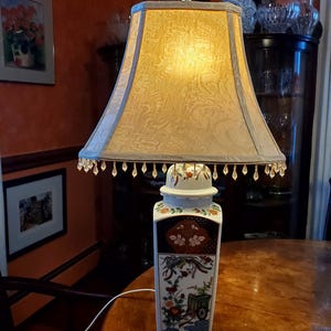 May include: A white porcelain lamp base with a floral and bird design. The lamp has a beige lampshade with a beaded fringe.