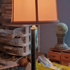 May include: A tall table lamp with a rectangular, clear glass base and a matching column. The lampshade is a light orange color with a brown trim. The lamp is on a wooden surface.