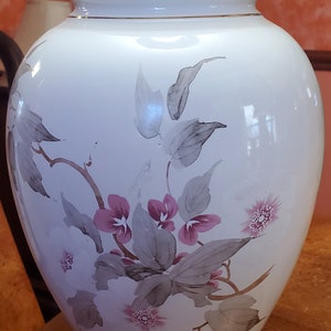 Peut inclure: Lampe de table en céramique blanche avec un motif floral composé de fleurs roses et de feuilles grises. La lampe a un abat-jour blanc et une base brune.