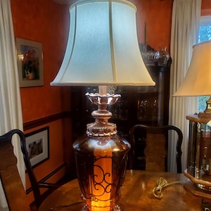 May include: A vintage amber glass table lamp with a white lampshade and a decorative metal base. The lamp is lit and casting a warm glow.