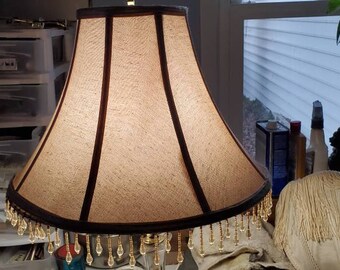 Puede incluir: Una pantalla de lámpara beige con ribete marrón oscuro y detalles verticales. La pantalla tiene una franja de cuentas a lo largo del borde inferior. La lámpara está en una habitación con una ventana y otros objetos decorativos.