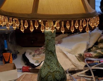 Puede incluir: Un jarrón de cerámica verde con una superficie texturizada se utiliza como base para una lámpara con una pantalla de tela beige. La pantalla tiene una franja de cuentas alrededor de la parte inferior.