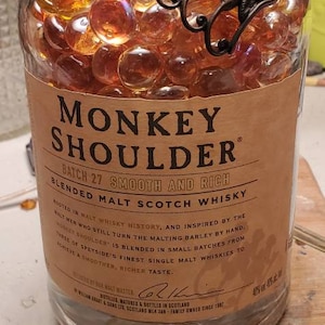 May include: A glass bottle of Monkey Shoulder blended malt scotch whisky. The bottle is filled with amber-colored glass beads and has a metal monkey shoulder ornament on the top. The label reads "Batch 27 Smooth and Rich" and includes information about the whisky's history and production.