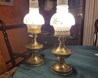May include: Two antique brass lamps with white glass shades. Each lamp has a round base, a central column, and a decorative top. The shades are decorated with a leaf design. The lamps are on a green tablecloth.
