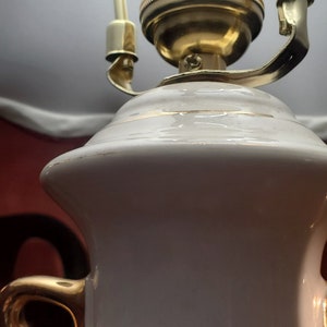 Può includere: Una base di lampada in ceramica bianca con dettagli dorati e un design floreale. La lampada ha un portalampada in metallo dorato con un interruttore e un paralume bianco.