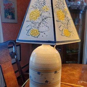 May include: A ceramic lamp base with a cream and brown glaze and blue floral accents. The lamp has a white lampshade with a blue trim and a floral design in yellow and blue.
