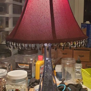 May include: A red lampshade with black trim and beaded fringe sits atop a blue and gold glass lamp base. The lamp is on a table with various jars and bottles.