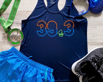 May include: A navy blue athletic tank top with the number "39.3" in orange and blue, paired with blue running shorts, black sneakers, a green medal, and blue sequined mouse ears. The items are arranged on a light wood-grain surface.