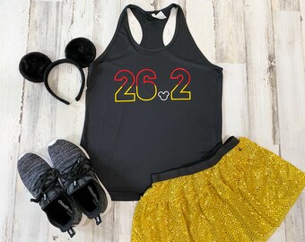 May include: Black athletic vest with the text "26.2" in red and yellow, a Mickey Mouse head, and a gold sequin skirt. Also included are black running trainers and black Mickey Mouse ears.
