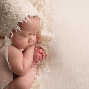 May include: A sleeping newborn baby wearing a white lace bonnet and a white lace ribbon. The baby is lying on a white textured background.
