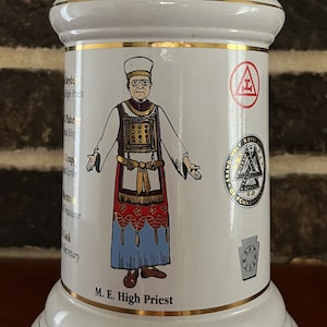 May include: White ceramic stein with a gold trim and a depiction of a man in a robe and a crown. The stein has the text "M.E. High Priest" and several Masonic symbols.
