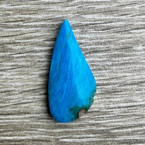 May include: A teardrop-shaped, polished gemstone in a vibrant turquoise hue. The stone has a smooth, glossy surface with visible natural patterns and a small area of earthy tones near the base. The gemstone is set against a textured, gray background.