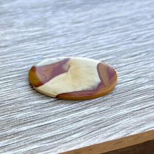 May include: Oval-shaped gemstone with a polished surface. The stone features a blend of colors, including cream, burgundy, and golden brown, creating a natural, earthy pattern. The gemstone is likely used for jewelry or decorative purposes.