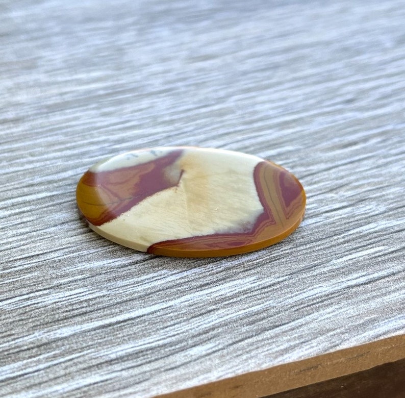 May include: Oval-shaped gemstone with a polished surface. The stone features a blend of colors, including cream, burgundy, and golden brown, creating a natural, earthy pattern. The gemstone is likely used for jewelry or decorative purposes.