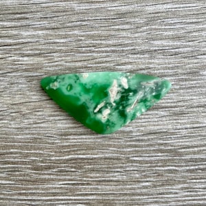 May include: A triangular, polished gemstone with a vibrant green hue, speckled with white and light gray inclusions. The stone is set against a textured, gray wooden surface, showcasing its natural beauty. Ideal for jewelry making or collecting.