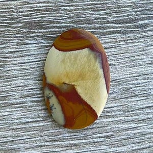 May include: Oval-shaped polished gemstone with a mix of colors. The stone features bands of brown, tan, and cream, with some areas of deep red. The gemstone is set against a textured, gray background.