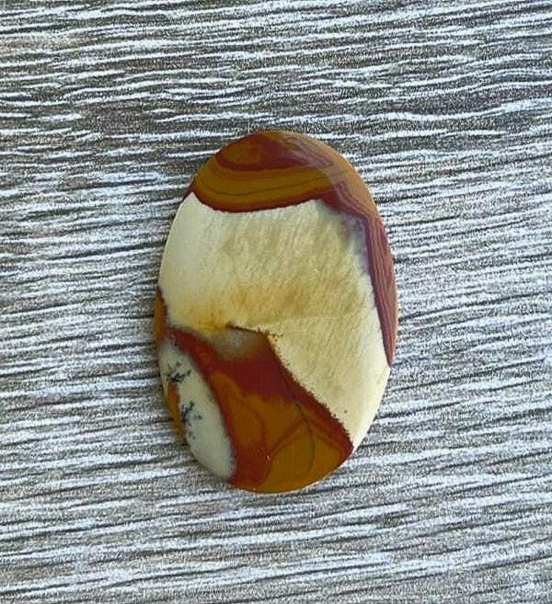May include: Oval-shaped polished gemstone with a mix of colors. The stone features bands of brown, tan, and cream, with some areas of deep red. The gemstone is set against a textured, gray background.
