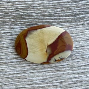 May include: Oval-shaped polished stone with a mix of colors. The stone features a cream-colored center, bordered by areas of deep red-brown and a lighter tan-brown. The stone's surface is smooth and reflective, with natural patterns.