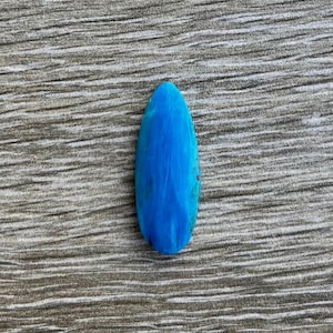May include: A vibrant, teardrop-shaped turquoise gemstone. The polished stone displays a bright blue hue with natural veining. The gemstone is set against a textured, gray background.