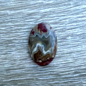 May include: An oval-shaped polished gemstone cabochon featuring a swirling pattern of deep red, earthy brown, creamy white, and muted gray tones. The stone has a smooth, glossy surface with distinct, flowing bands of color throughout the piece.