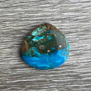 May include: A polished, teardrop-shaped gemstone with a striking color palette. The stone features a vibrant turquoise blue base, transitioning to areas of deep teal and brown. The surface has a textured appearance, with natural variations in color and pattern.