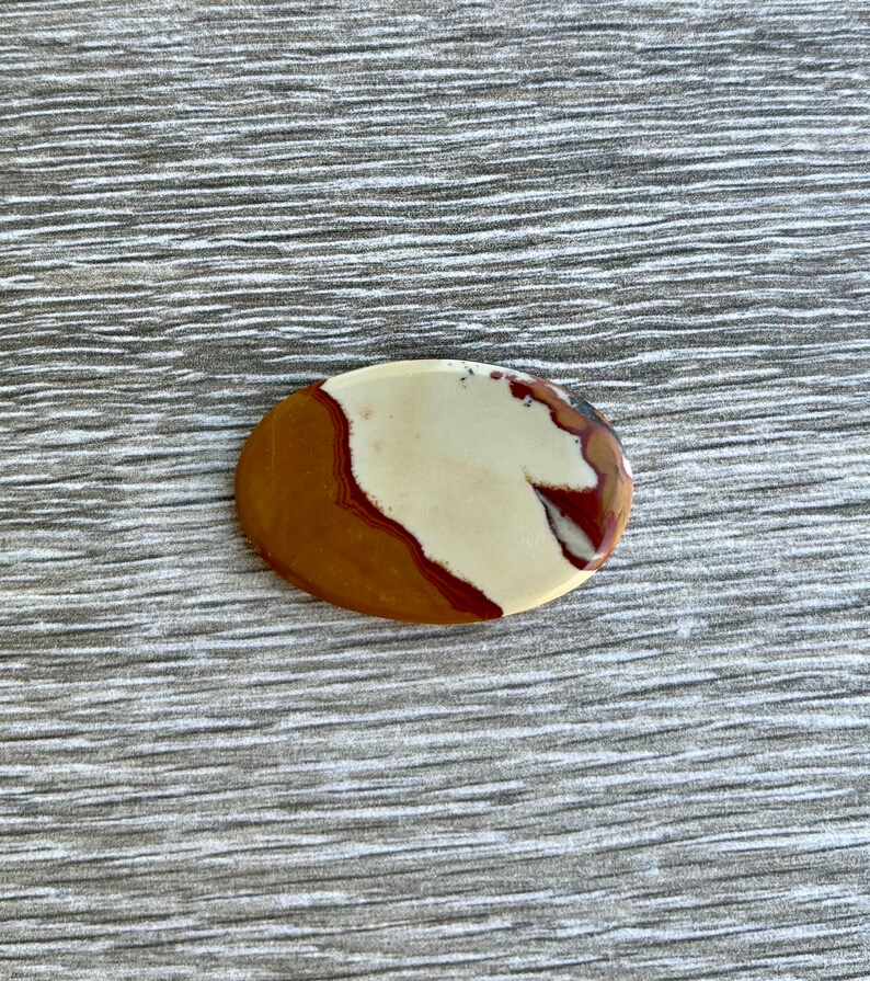 May include: An oval-shaped polished stone with a mix of colors. The stone features a base of tan and brown, with a creamy white section and streaks of deep red. The stone's surface is smooth and reflective, suggesting it has been polished.