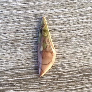 May include: A polished, teardrop-shaped gemstone with a mix of colors. The stone features shades of green, brown, and pink, with a glossy finish. The gemstone is set against a light gray, wood-grain surface.