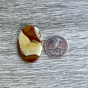 May include: An oval-shaped gemstone with a mix of tan, brown, and cream colors. The stone is next to a 2018 Block Island quarter for size comparison. The gemstone has a polished surface and a natural, earthy appearance.