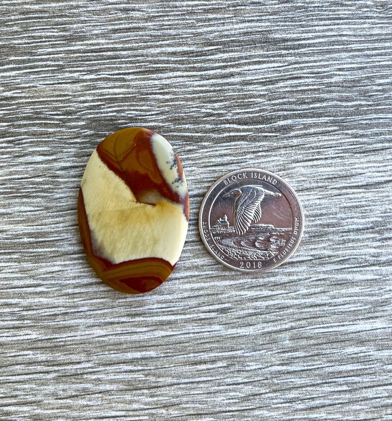 May include: An oval-shaped gemstone with a mix of tan, brown, and cream colors. The stone is next to a 2018 Block Island quarter for size comparison. The gemstone has a polished surface and a natural, earthy appearance.