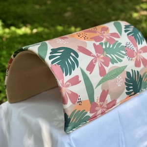 Puede incluir: Una cama para gatos curvada, beige y blanca, con un estampado tropical con flores rosas y naranjas y hojas verdes.