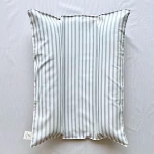 May include: A rectangular pillow with a white background and vertical stripes in a muted green color. The pillow has a tag attached to the bottom left corner. The pillow is likely for home decor.