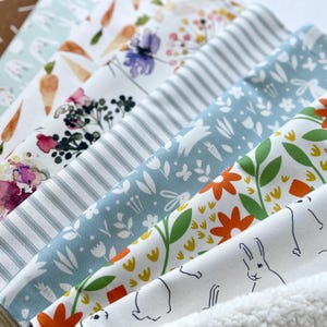 May include: A selection of patterned fabrics featuring floral, carrot, and bunny designs. Colors include white, blue, orange, and green. The fabrics are arranged diagonally, showcasing various patterns and textures, suitable for crafting and home decor projects.