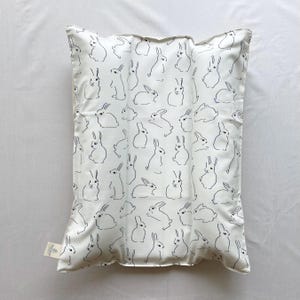 May include: White pillow with a repeating pattern of navy blue rabbit illustrations. The pillow has a soft, satin-like texture and a tag on the lower left corner. The rabbits are depicted in various poses.