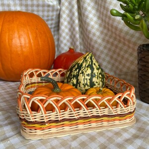 May include: A woven basket with a brown, orange and yellow design holds three small pumpkins. One pumpkin is green with white stripes, one is orange and one is a small, round, orange pumpkin.