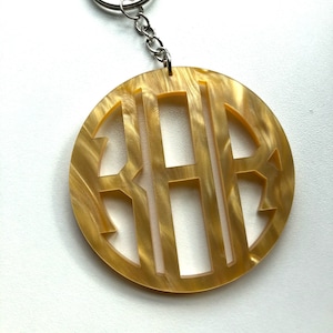 May include: A gold-toned monogram keychain with a circular design and a silver key ring. The monogram is cut out of the circle, revealing the gold-toned background.
