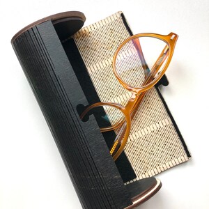 K&ouml;nnte beinhalten: Ein schwarzes zylindrisches Brillenetui mit einem Holzrand und einem braunen Innenraum. Das Etui ist ge&ouml;ffnet und ein Paar runde, bernsteinfarbene Brillen mit klaren Gl&auml;sern liegt darin.