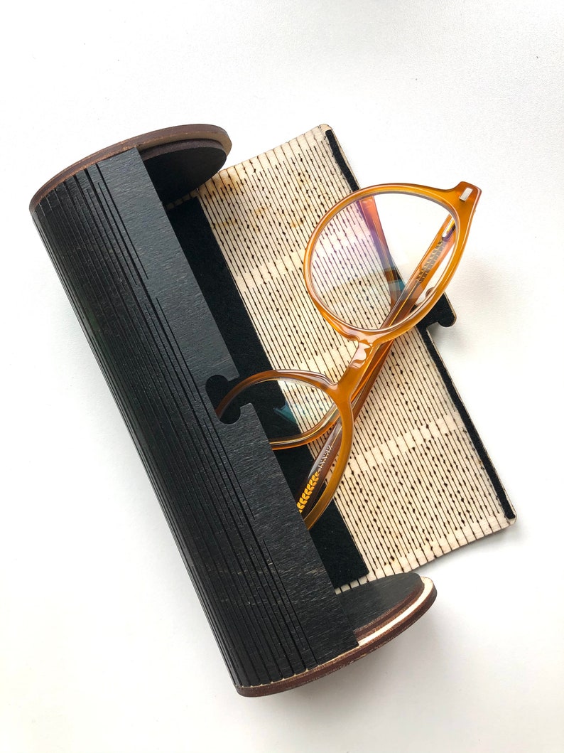 K&ouml;nnte beinhalten: Ein schwarzes zylindrisches Brillenetui mit einem Holzrand und einem braunen Innenraum. Das Etui ist ge&ouml;ffnet und ein Paar runde, bernsteinfarbene Brillen mit klaren Gl&auml;sern liegt darin.