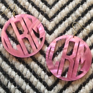 May include: A pair of pink monogram earrings with a marbled effect. The earrings are made of a lightweight material and have a gold-tone hook closure.