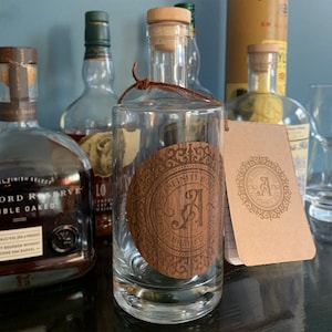 May include: A clear glass bottle with a cork stopper and a wooden label that says "Infinity Bottle Established 1962". The bottle is sitting on a dark surface with other bottles in the background.