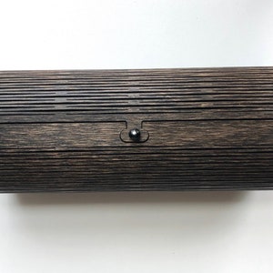 K&ouml;nnte beinhalten: Eine zylindrische Holzkiste mit dunkelbrauner Oberfl&auml;che und einem schwarzen Verschluss. Die Kiste hat ein gerilltes Muster auf der Au&szlig;enseite.