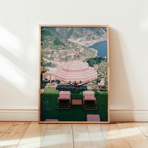 Può includere: Un ombrello a strisce rosa e bianche con due sdraio rosa su una collina erbosa che domina un mare blu e una collina verde.