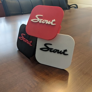 May include: Three square-shaped plaques with the word "Scout" written in a stylized font. The plaques are red, black, and white.