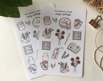 May include: Two sheets of stickers with a cozy cottage theme. The stickers feature illustrations of a cup of coffee, a pie, a book, an envelope, flowers, and a mountain landscape. The text "The Aesthetics Studio cozy cottage" is printed on each sheet.