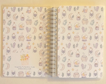 Può includere: Un quaderno a spirale bianco con un design fantasioso che presenta lune, cristalli, bottiglie di pozioni e un calderone. La copertina ha il testo "Designed by The Aesthetics Studio @theaestheticstudio".