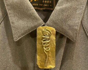 Op de afbeelding: Een gouden bolo tie met een gedraaid draadontwerp. De bolo tie wordt gedragen op een grijs shirt met een label waarop staat "The Howler B-R-O-S Need The Call".