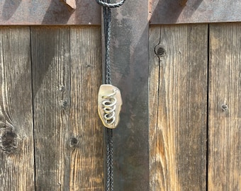 Op de afbeelding: Een zwarte bolo tie met een zilveren metalen hanger met een gekruld ontwerp. De stropdas hangt aan een roestige metalen poort.