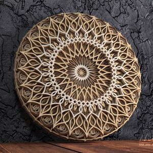 K&ouml;nnte beinhalten: Ein mehrschichtiges Mandala-Wandkunstwerk aus Holz. Das kreisf&ouml;rmige Design weist komplizierte Muster in Beige- und Wei&szlig;t&ouml;nen auf, wodurch ein dreidimensionaler Effekt entsteht. Das Kunstwerk wird vor einem dunklen, strukturierten Hintergrund pr&auml;sentiert.